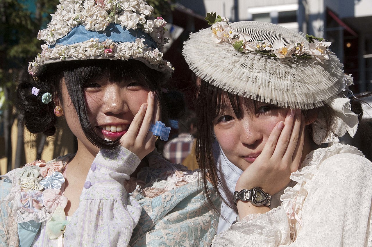 Two young women in Harajuku wearing layered pastel lace tops with flower-trimmed hats and ribbon hair accessories