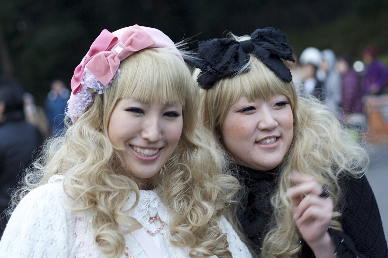 Two women in Harajuku with blonde curled hair, one in a pink velvet bow beret and cream knit, one in a black lace bow and black top