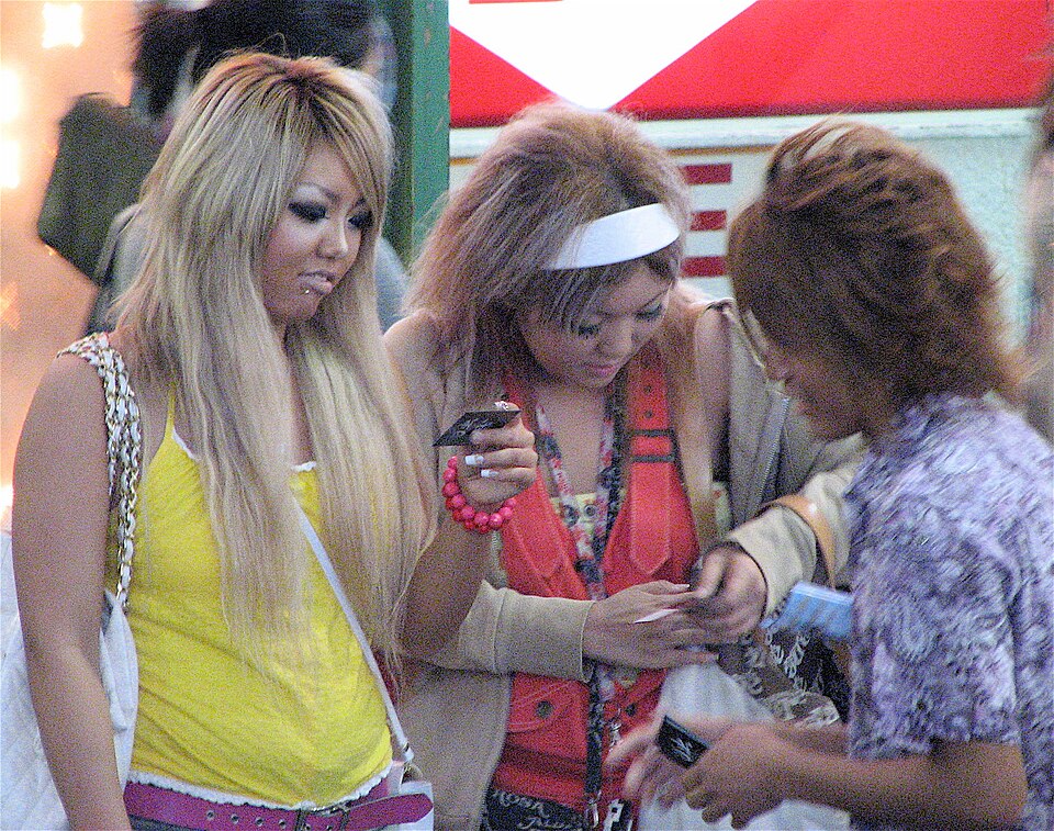 Three young women outdoors with lightened hair looking at a flip phone, wearing a yellow tank top, red vest, and purple print top