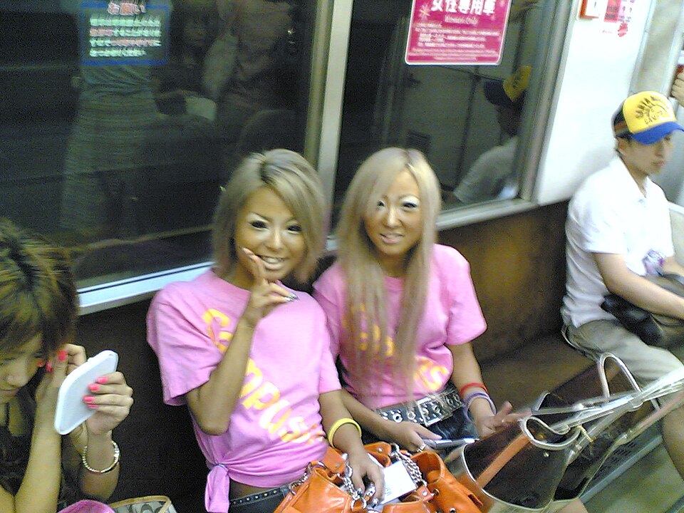 Two Japanese women in ganguro gyaru style with bleached hair, pink logo tees, and studded belts on a train