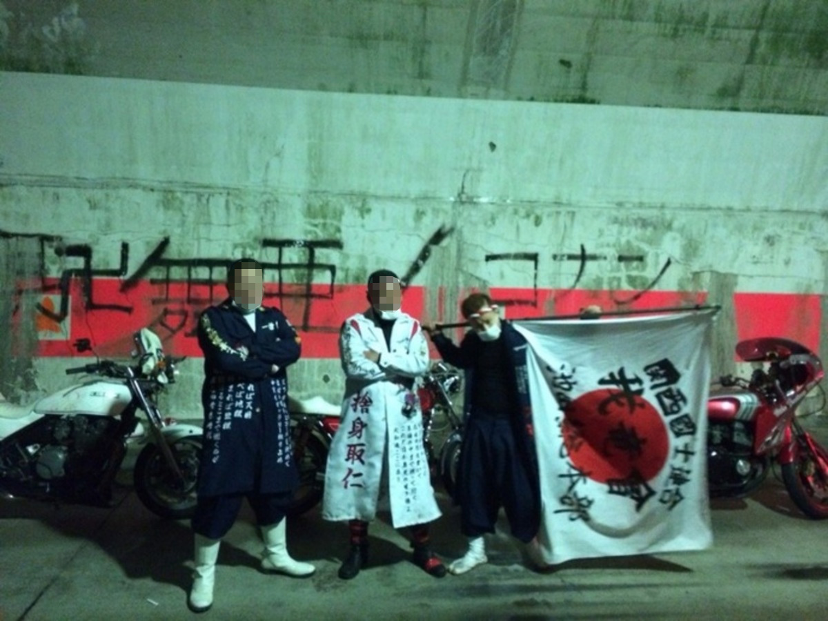 Three bosozoku members in long embroidered coats holding a rising sun banner beside parked bikes under a night underpass