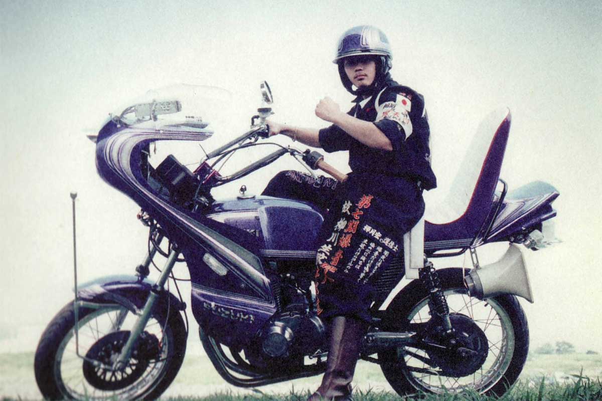 Vintage photo of a bosozoku rider in an embroidered jacket seated on a purple bike with a rocket cowl and tiered pillion seat