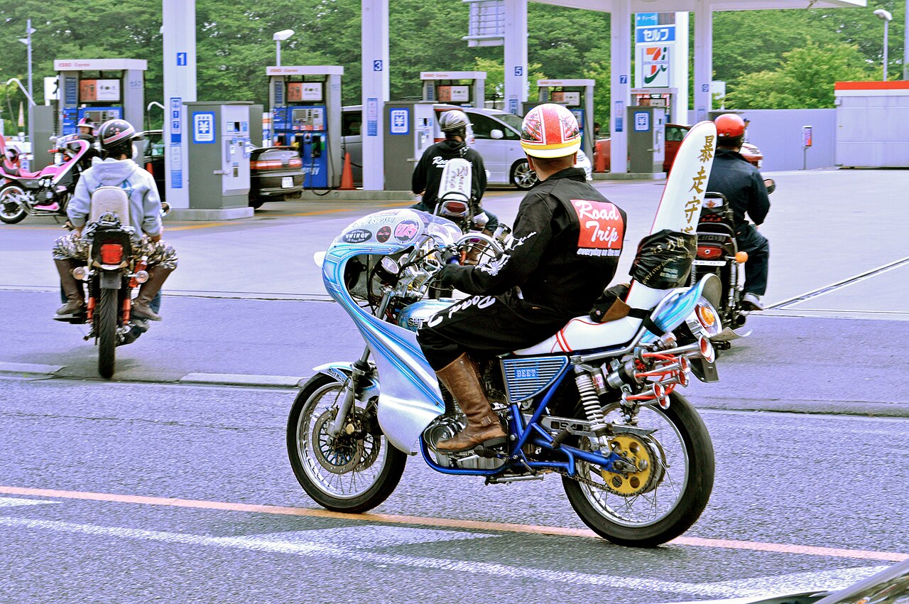 Bosozoku riders on customised motorcycles with tall rocket cowls and kanji jackets pulling out of a Japanese petrol station