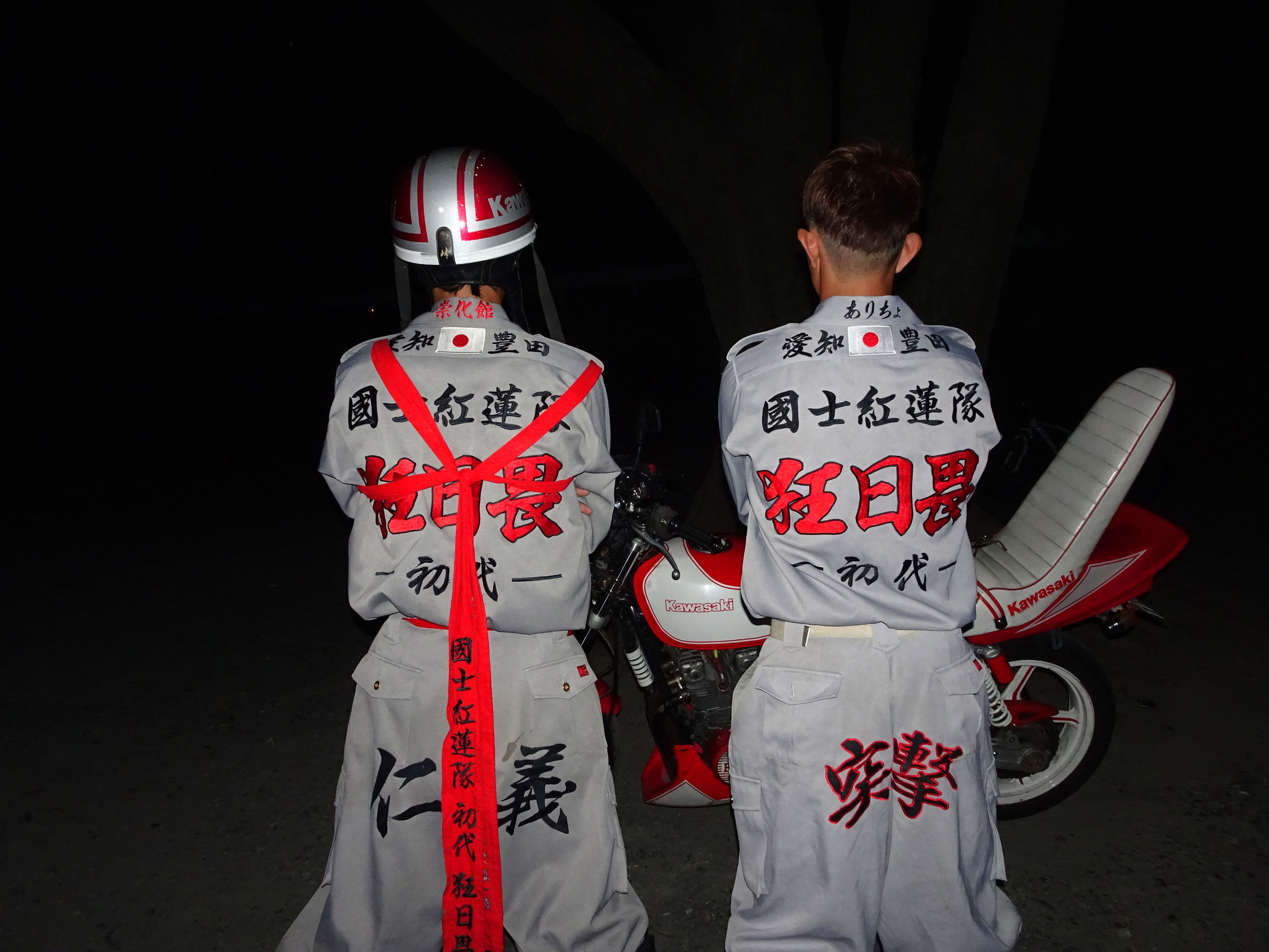 Two bosozoku members photographed from behind in grey tokko-fuku jumpsuits embroidered with red and black kanji and rising sun patches