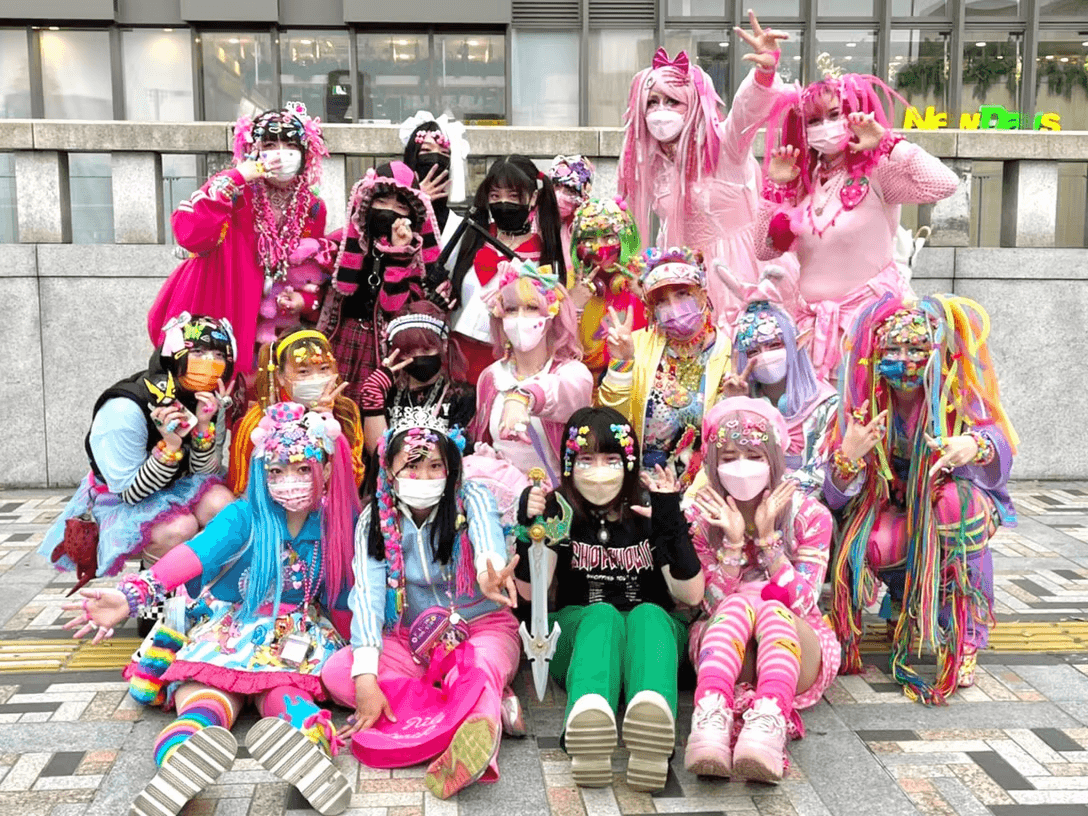 Large group of decora wearers posed together outdoors in pink, pastel and dark coordinates with rainbow yarn falls, layered hair clips, striped socks and patterned face masks