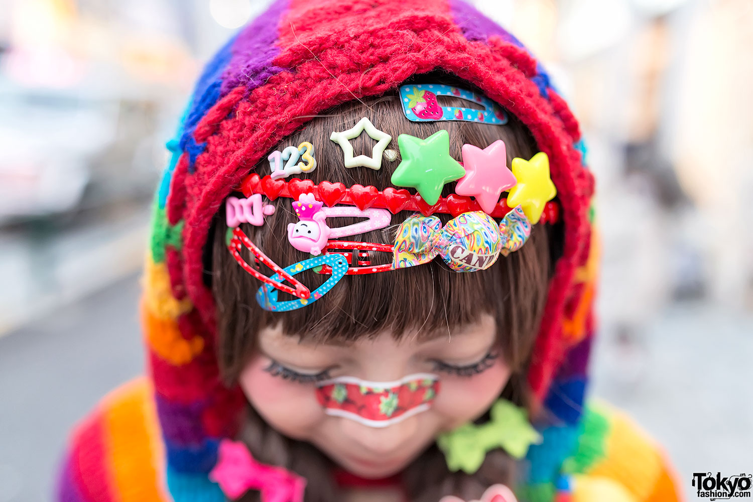 Girl in a rainbow knit hood with rows of star, heart and character hair clips pinned across her bangs and a patterned band-aid worn over the bridge of her nose
