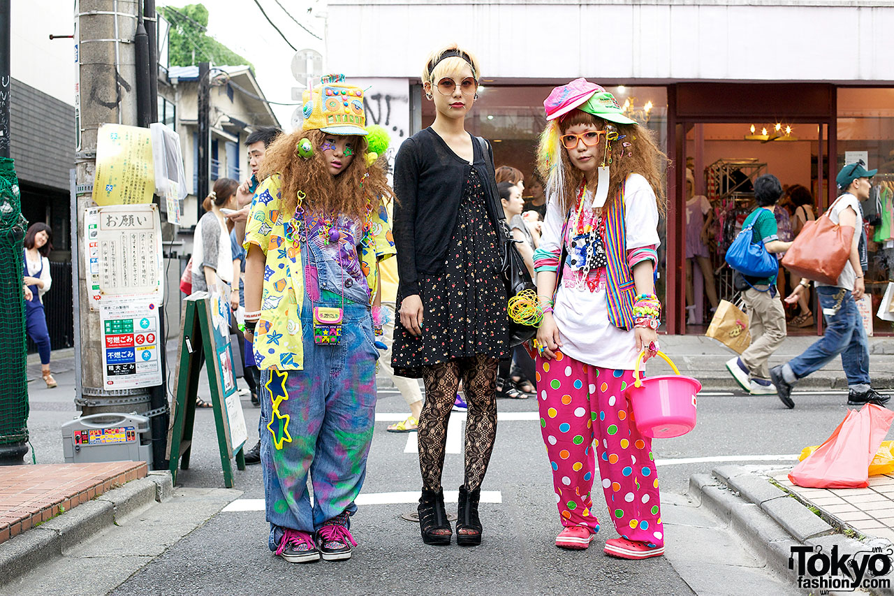 Three young people photographed on a Harajuku street, two in bright decora style outfits with printed caps, tie-dye overalls and polka dot trousers, one in plain black