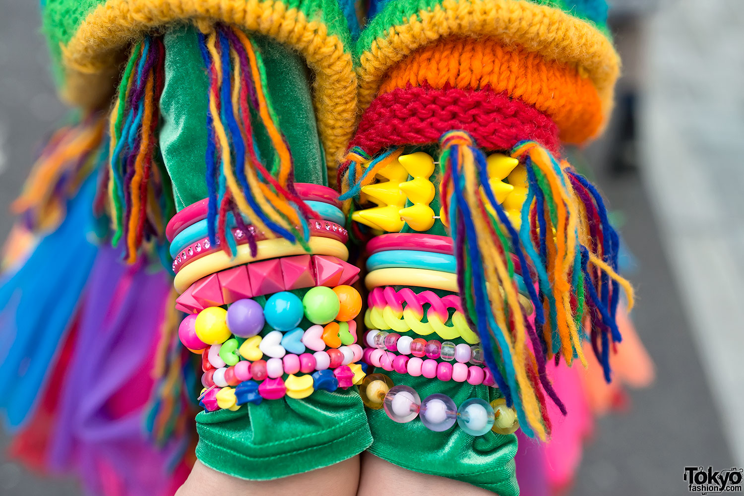 Close-up of two forearms almost completely covered by stacked decora bracelets in bright plastic beads, bangles and rainbow chain links, worn under a rainbow knit sleeve
