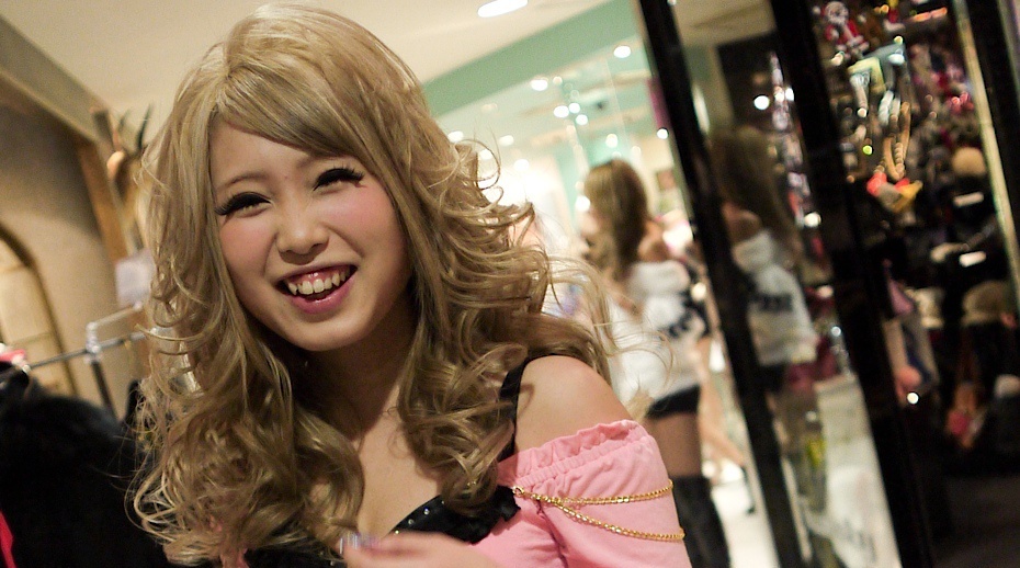 Young woman with curled bleached hair, heavy false eyelashes, and a pink off-shoulder top with chain detail inside a Shibuya boutique
