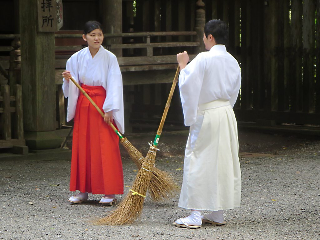 Shrine maiden in bright red hakama and a Shinto priest in pale hakama sweeping a shrine yard