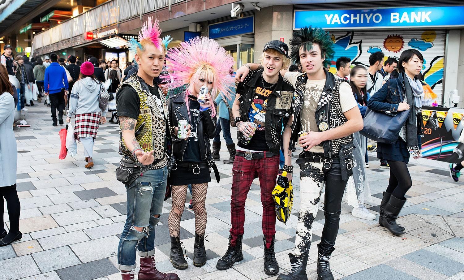 Four punks in Harajuku wearing studded leather vests and plaid trousers, with pink, teal, and two-tone mohawks, drinking canned drinks on a shopping street