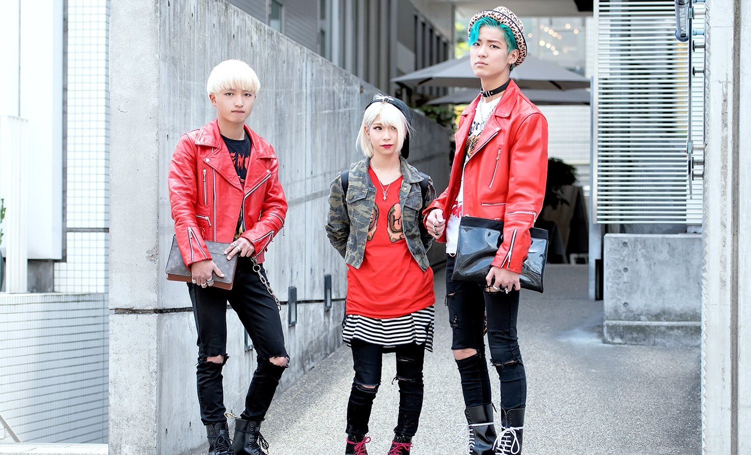 Two people in red leather biker jackets, one with bleached blonde hair and one with teal green hair, standing with a friend in a camo denim jacket over a red tee, all in black ripped jeans and boots