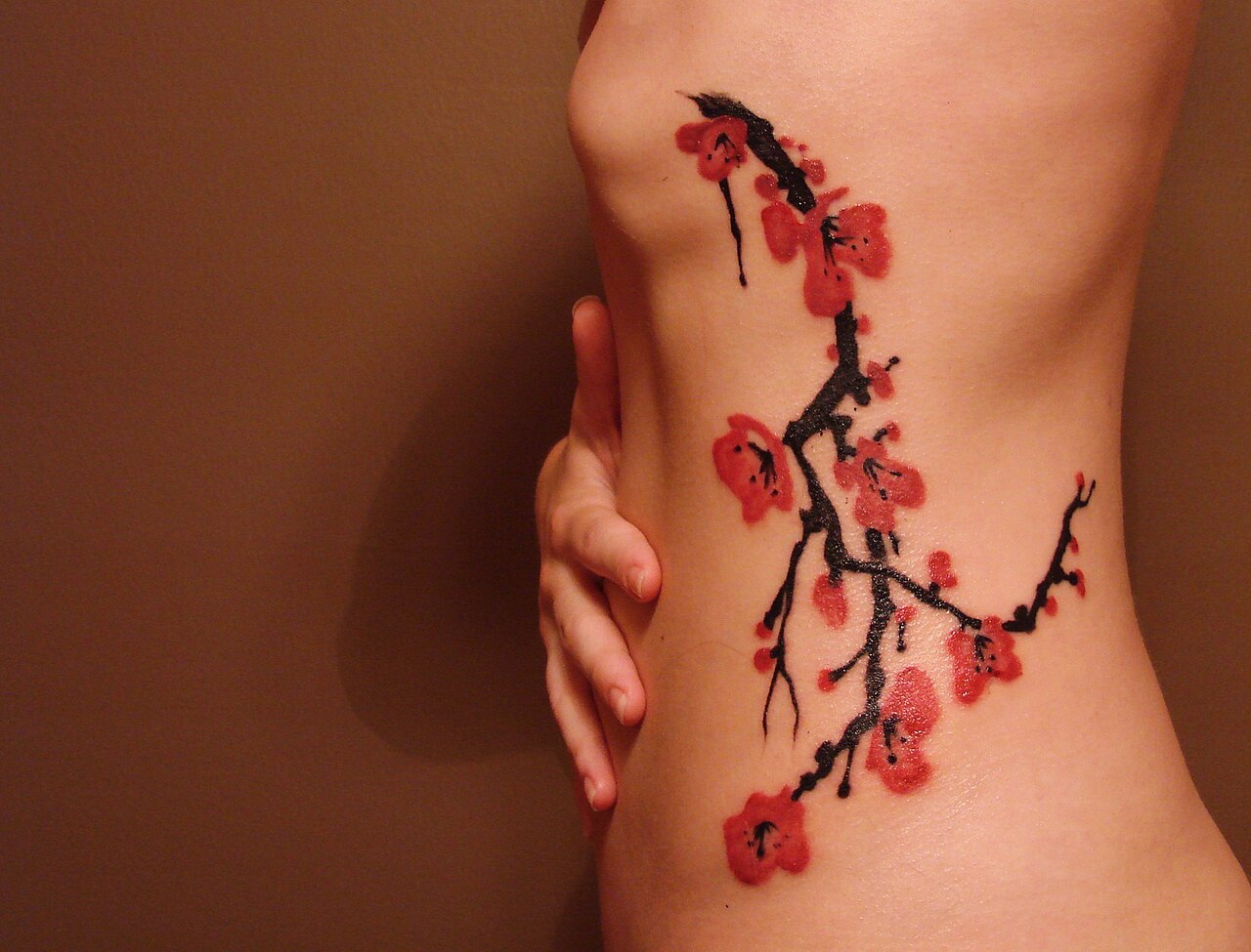 Cherry blossom branch tattoo in red and black ink running along a person's ribs and hip