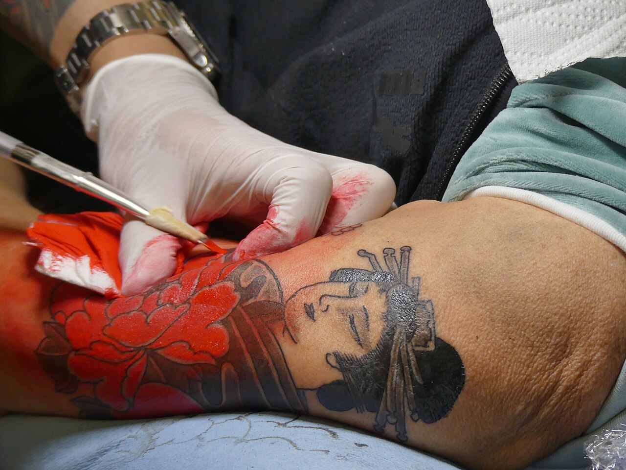 Tattoo artist hand-poking a red peony and figure in traditional Japanese style using tebori tools
