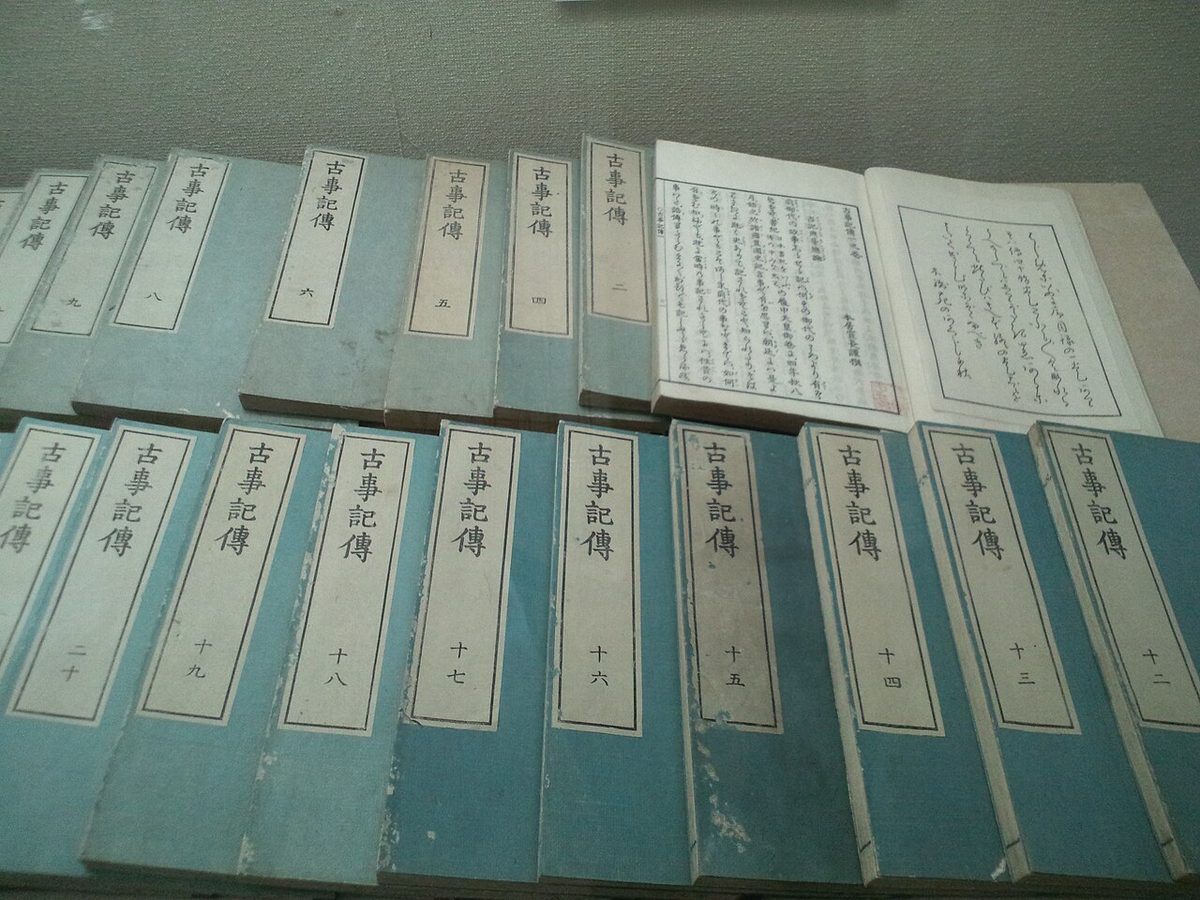 Rows of blue bound Kojiki den volumes with Japanese title labels, one open to handwritten pages