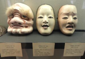Three carved Japanese Noh and Kagura masks displayed side by side in a museum case