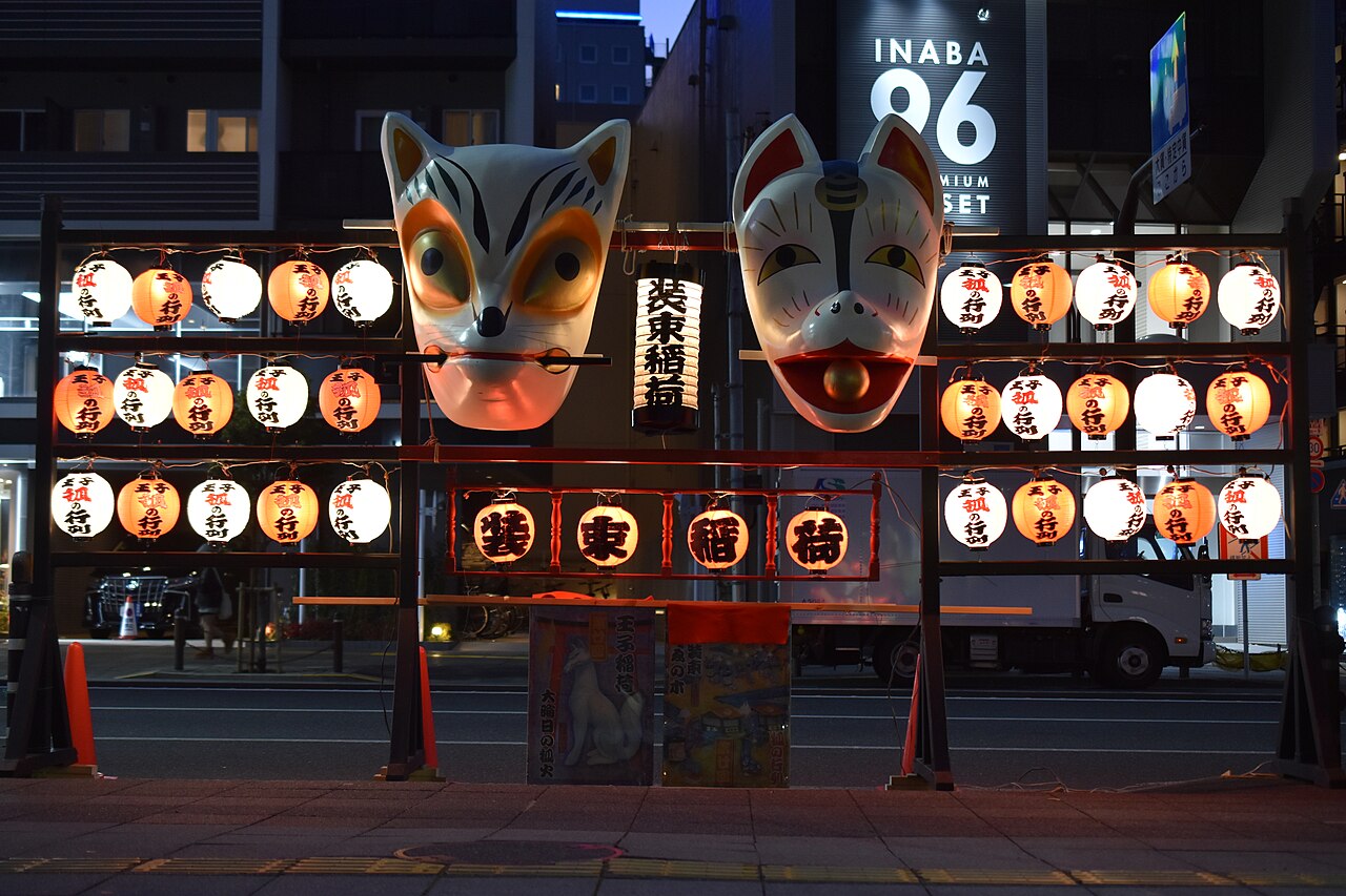 Two large kitsune fox masks mounted above rows of glowing paper lanterns at a night shrine procession