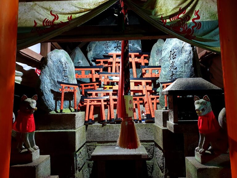 Two stone fox guardians in red bibs flanking a miniature torii altar at a Fushimi Inari sub-shrine