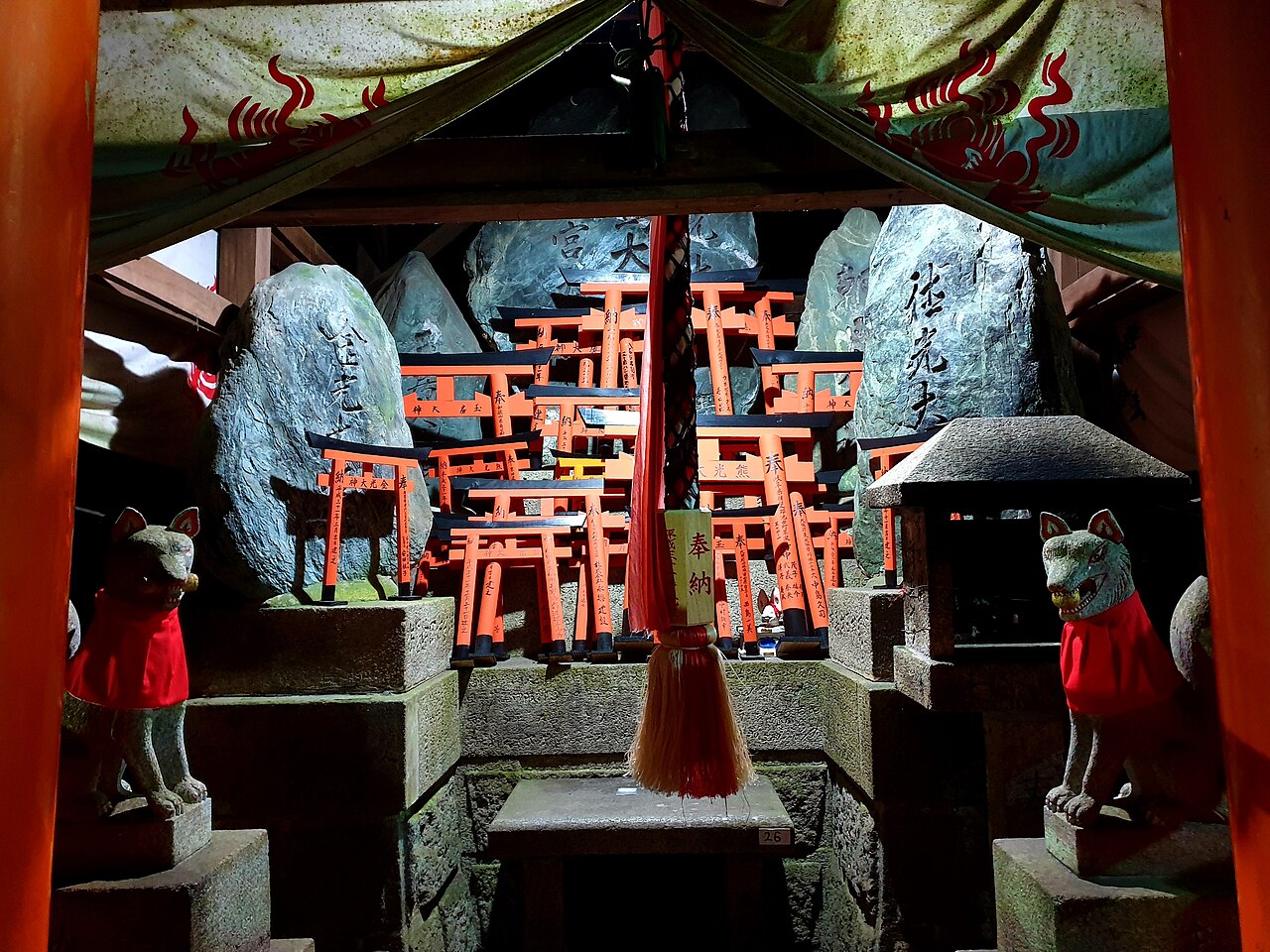 Two stone fox guardians in red bibs flanking a miniature torii altar at a Fushimi Inari sub-shrine