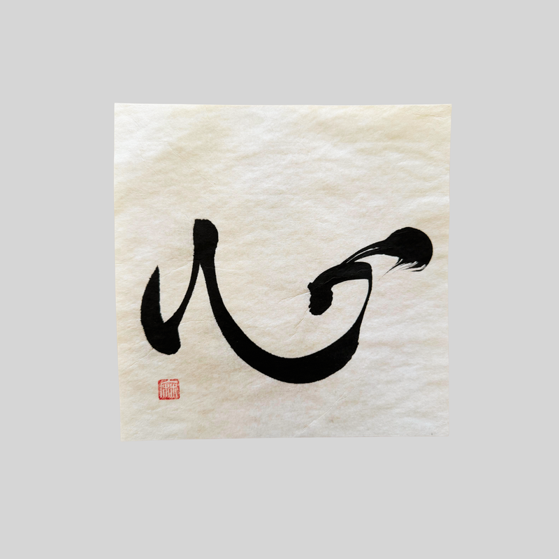 The kanji 心 (kokoro) brushed in black ink on cream washi paper with a red seal in the corner