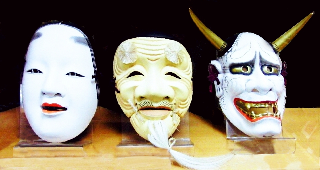 Three carved noh masks on stands: a pale young woman, an old man, and a white horned hannya with gold fangs