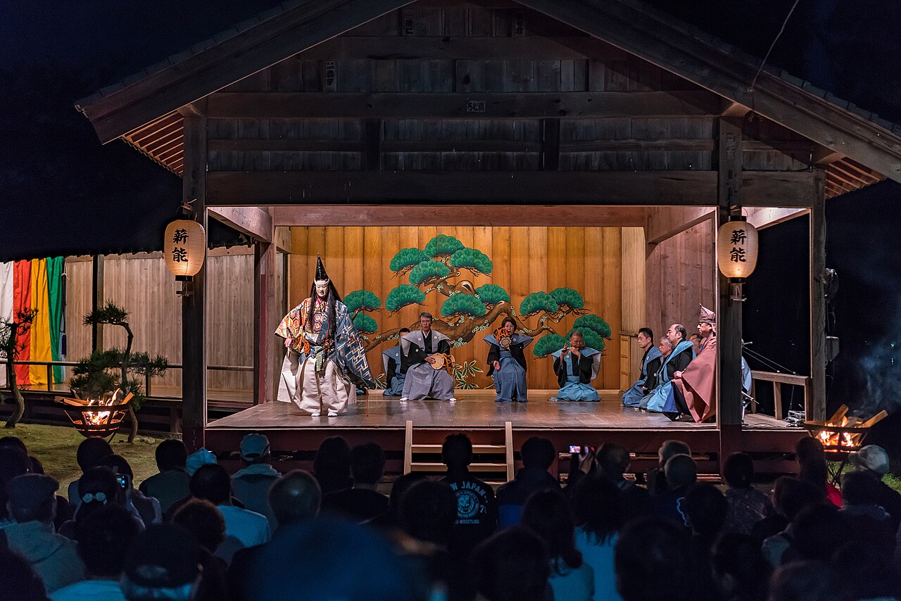 Masked noh actor performing on an open-air wooden stage lit by firelight in front of a seated crowd