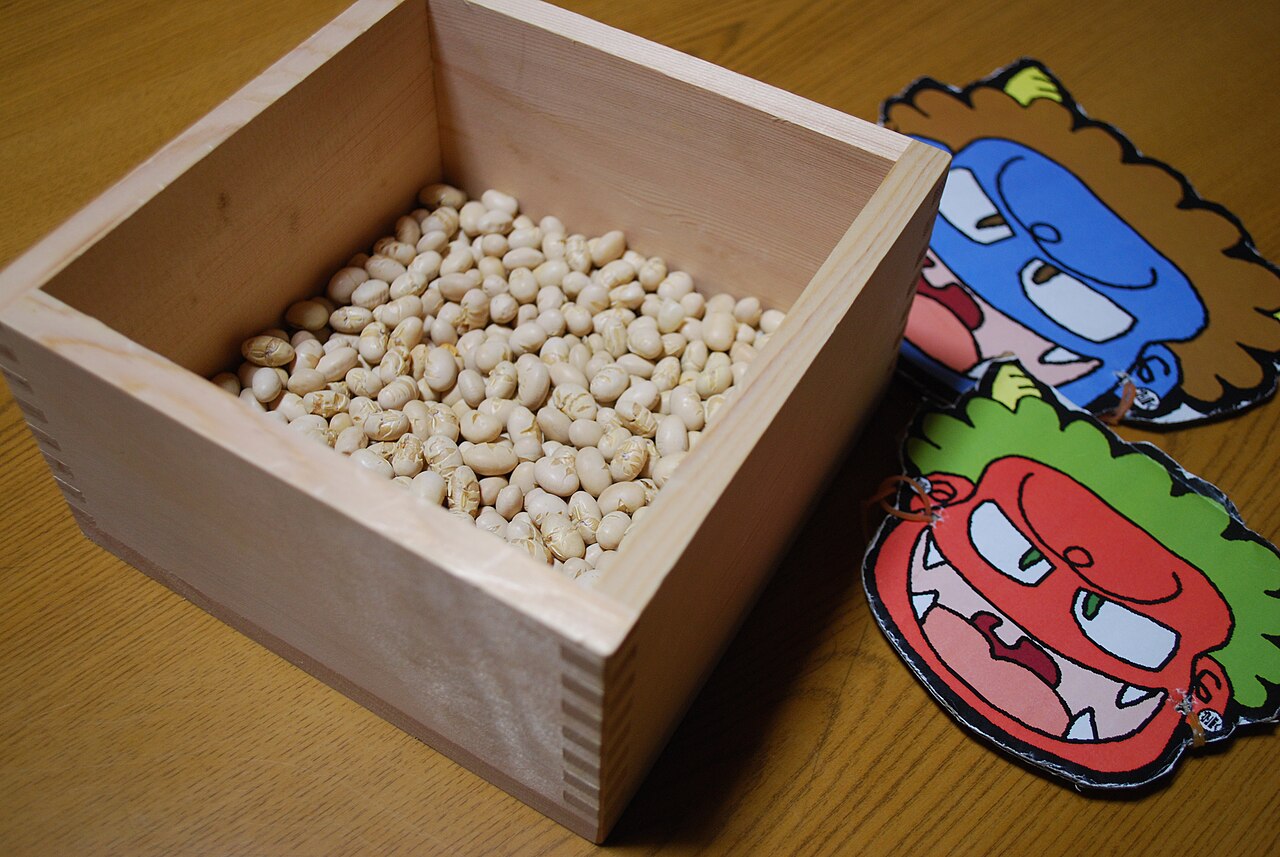 Wooden masu box of roasted soybeans beside red and blue paper oni masks for the Setsubun bean-throwing ritual
