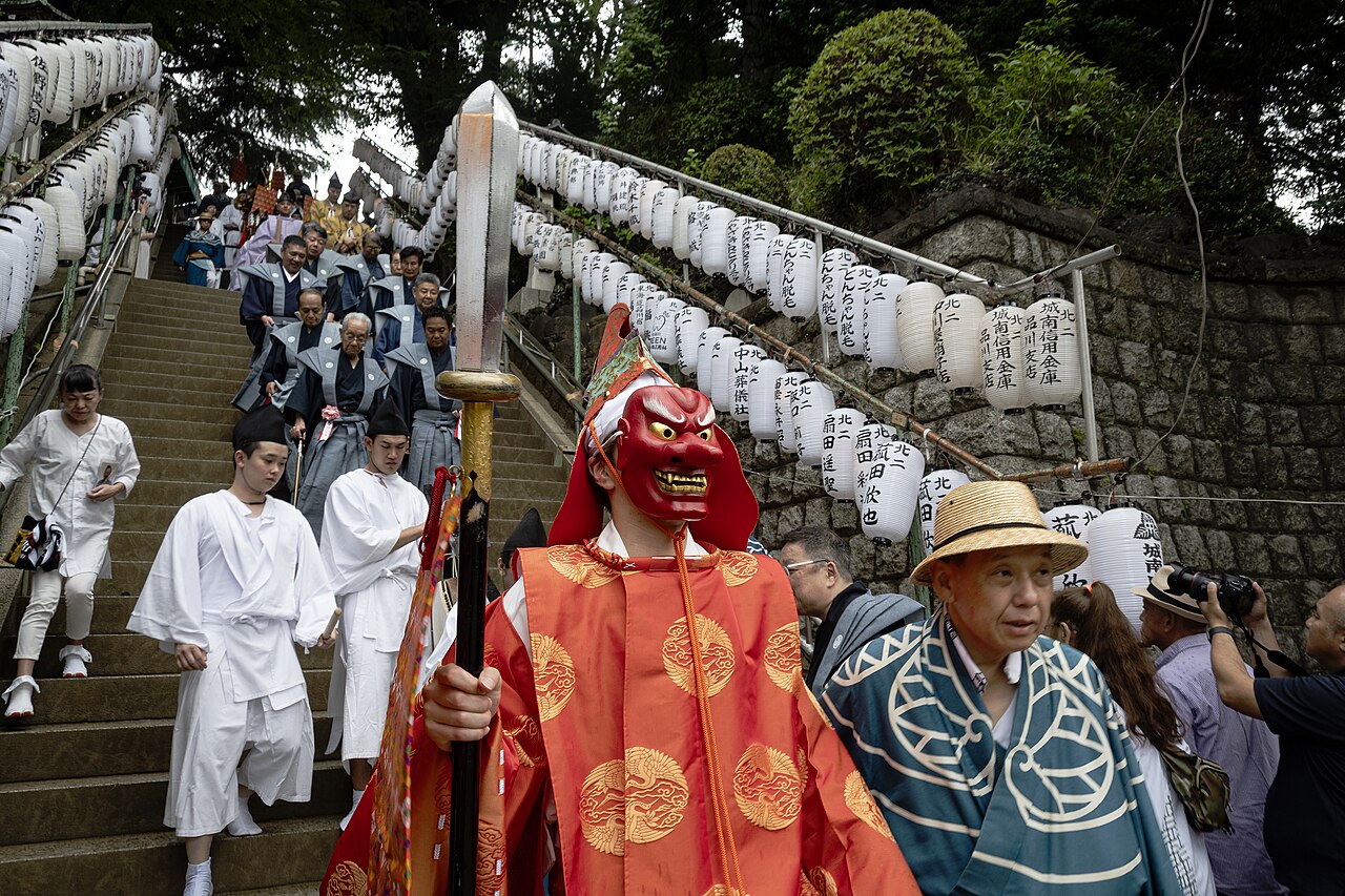 Performer in a red tengu mask with a long nose carrying a sword down shrine steps hung with paper lanterns