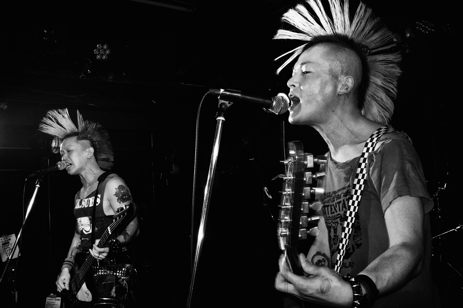 Black and white photo of two Japanese punk musicians with bleached mohawks performing live on stage, one playing bass and one playing guitar
