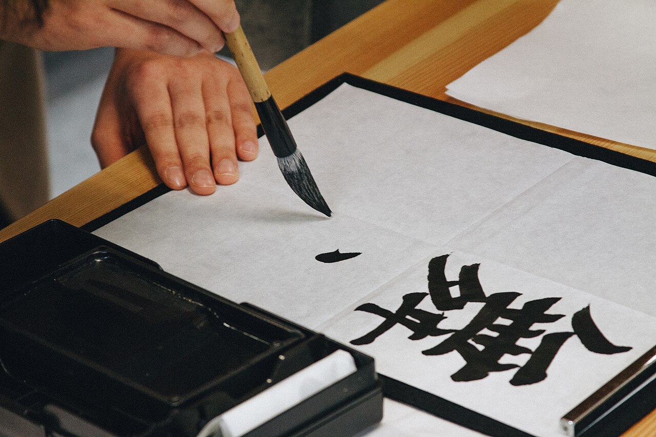 Hand holding a calligraphy brush over rice paper with a freshly inked kanji character beside an ink tray