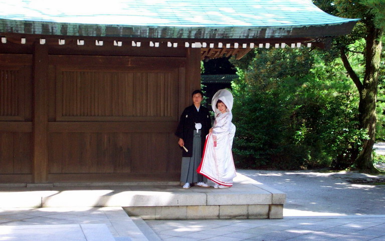 Bride in a white shiromuku and groom in a black crested montsuki hakama standing at a Japanese shrine