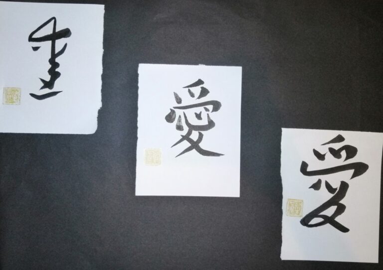 Three sheets of paper on a dark surface, each brushed in black ink with the kanji 愛, the Japanese symbol for love