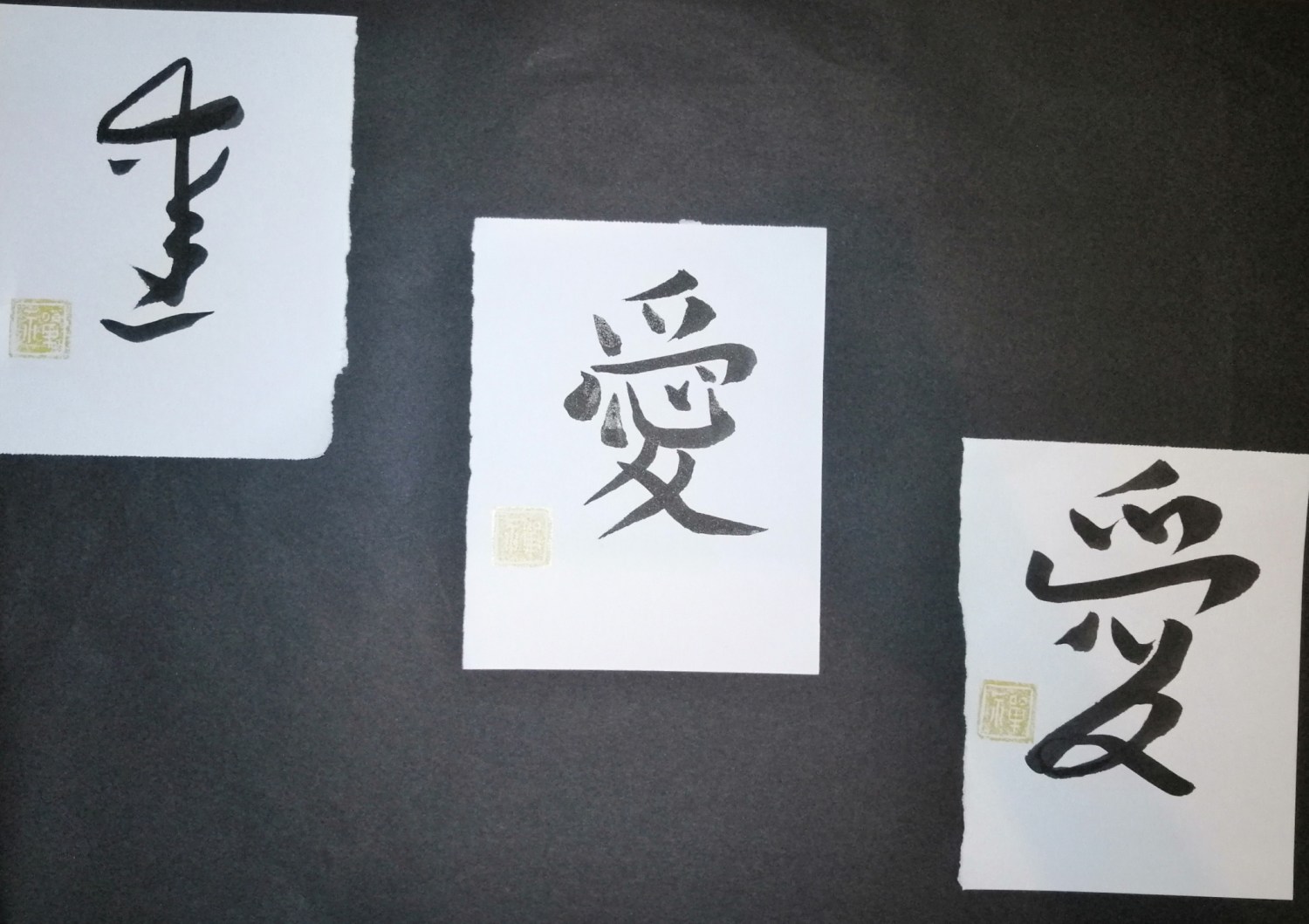 Three sheets of paper on a dark surface, each brushed in black ink with the kanji 愛, the Japanese symbol for love