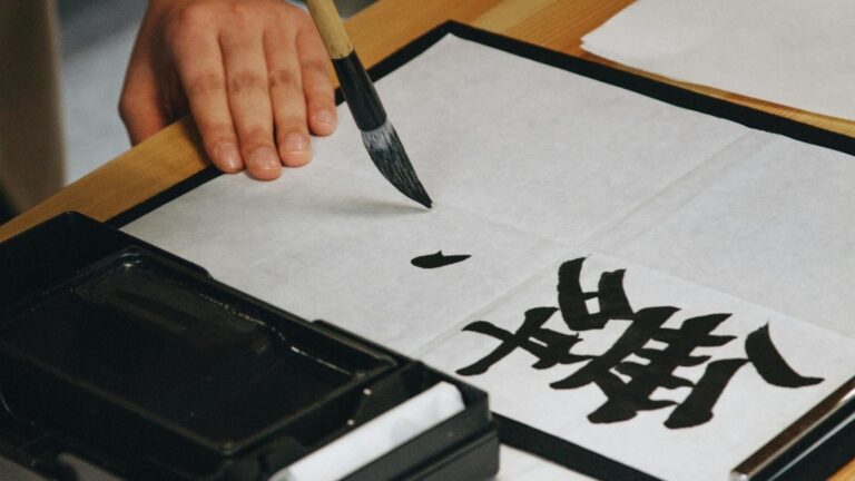 A hand holding a calligraphy brush over paper beside a freshly brushed black kanji character, with an inkstone in the foreground