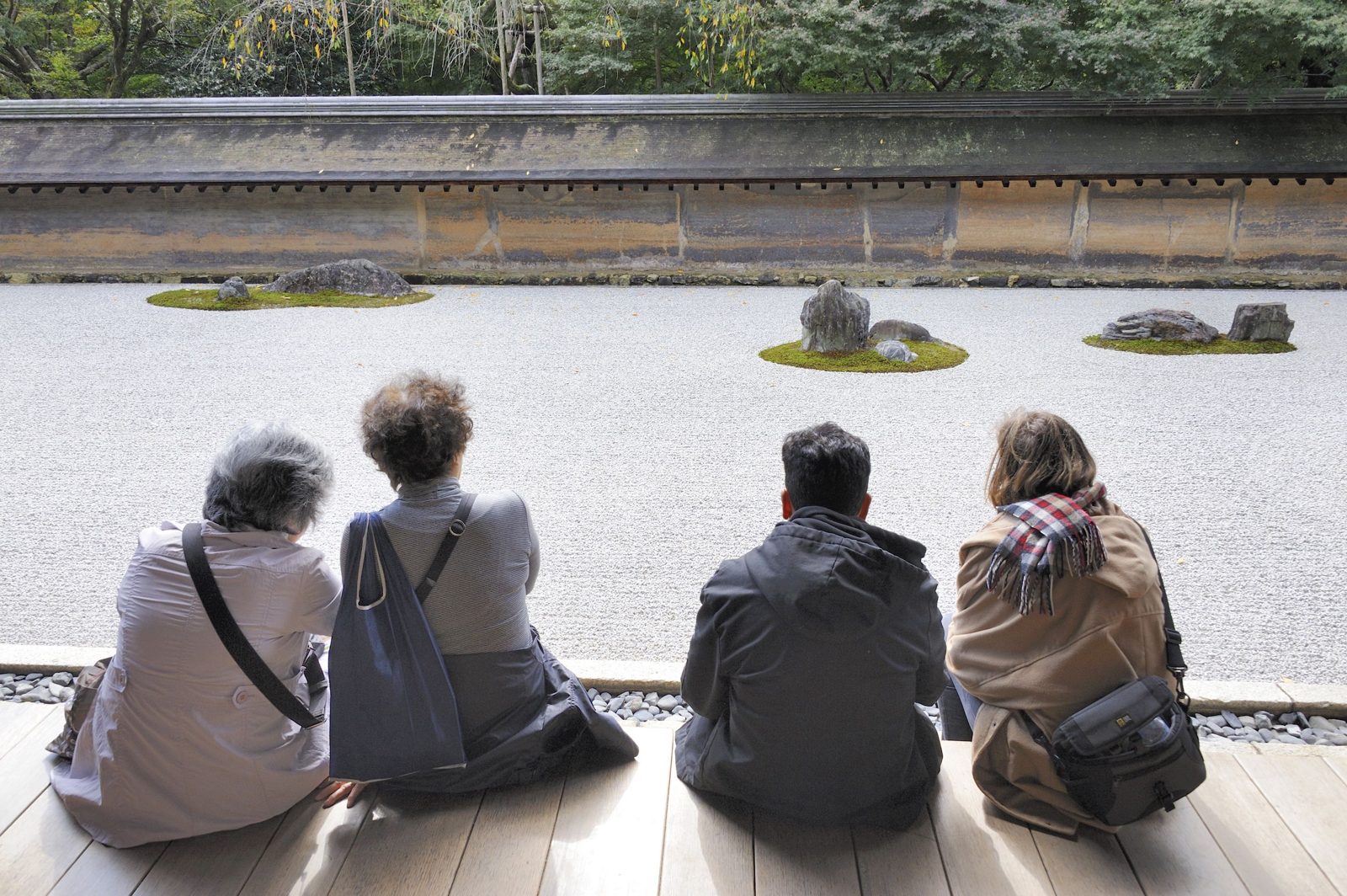 Four visitors sitting on a wooden veranda looking out at the raked gravel and moss-ringed rocks of the Ryoanji zen garden in Kyoto