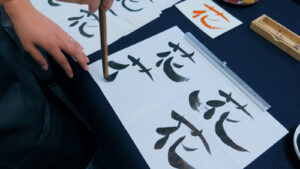 A hand brushing the kanji 花 in black ink on white paper with a bamboo calligraphy brush