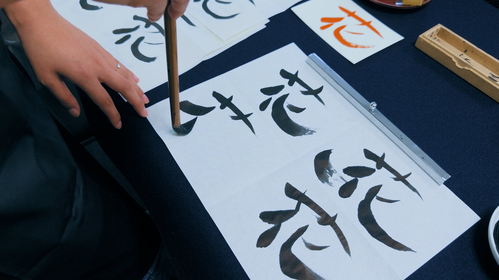 A hand brushing the kanji 花 in black ink on white paper with a bamboo calligraphy brush