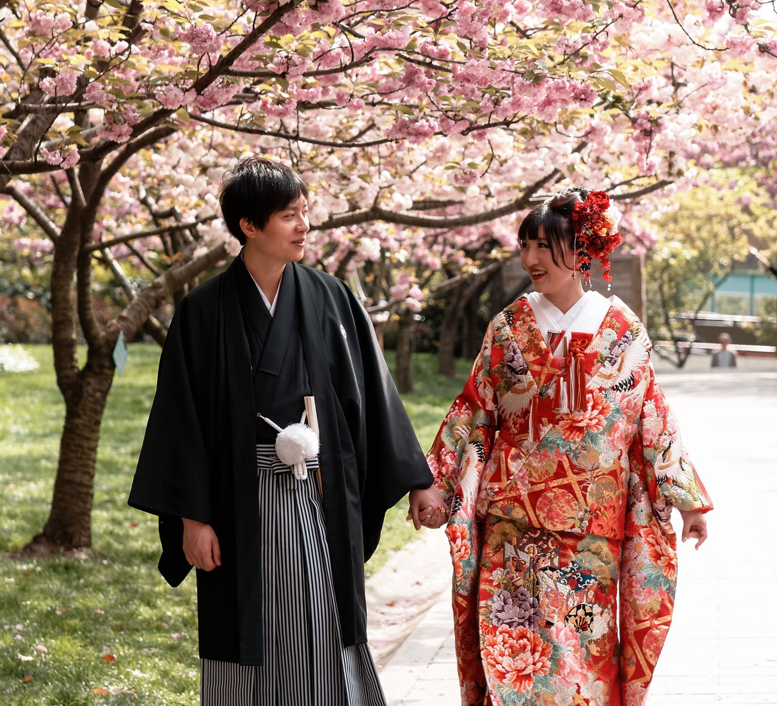 Bride in an orange iro-uchikake with white crane embroidery walking beside a groom in black montsuki and striped hakama