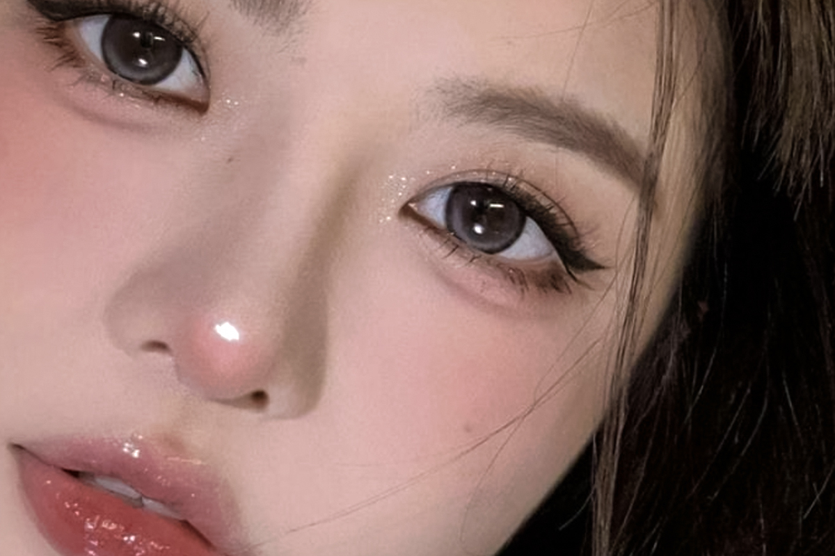 Close up of a pale made up face with soft light brows, thin black liner that drops at the outer corner instead of flicking up, cool pink shimmer under the eye and a glossy pink lip