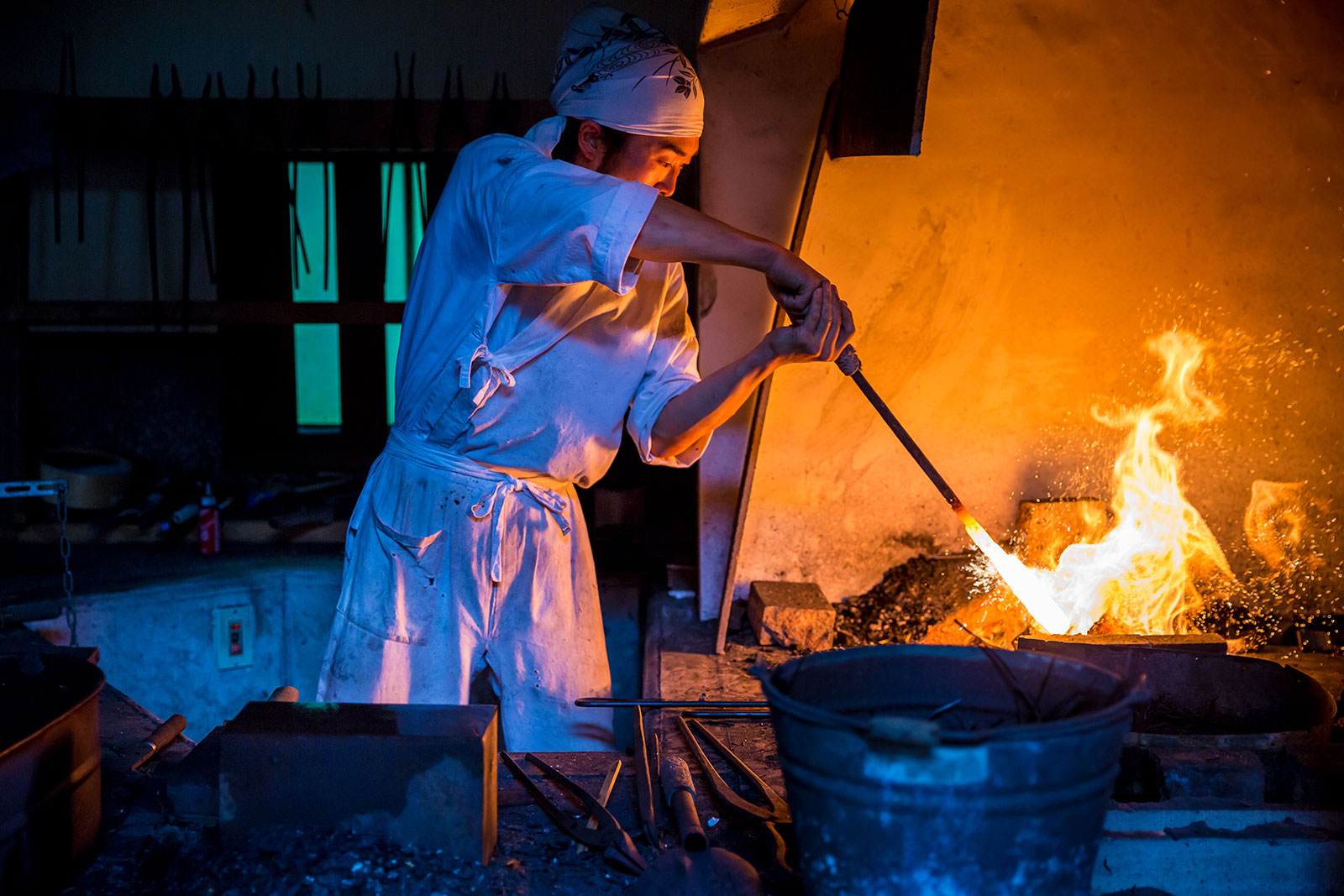 Japanese swordsmith in white robes holding glowing steel over an open forge fire, sparks flying in a dark workshop