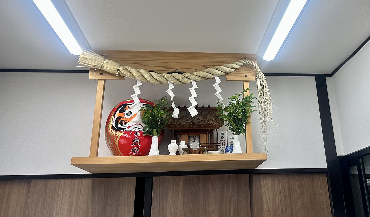 Household kamidana shelf with a straw shimenawa rope, folded paper streamers, and a red Daruma doll