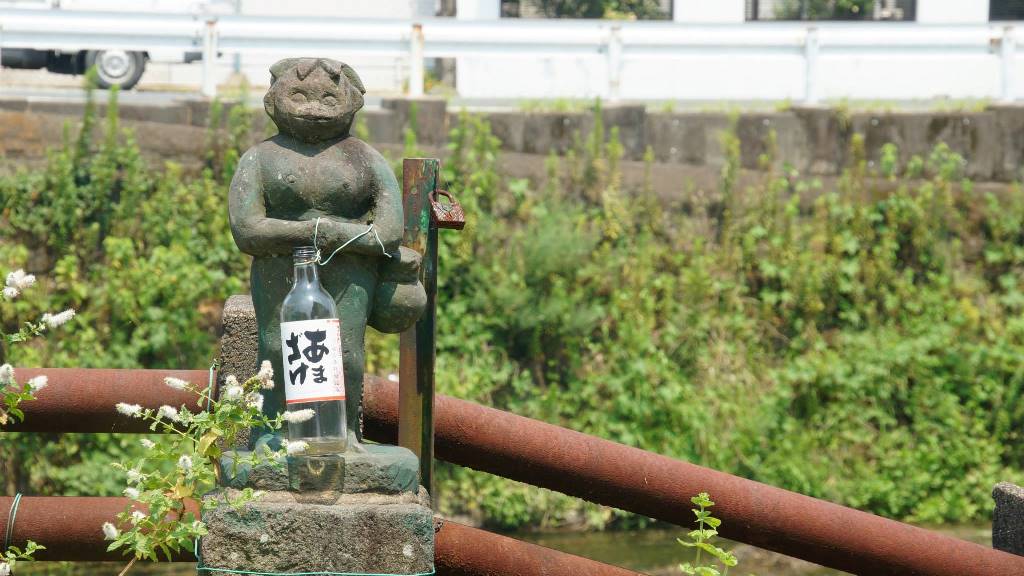 Small stone kappa statue on a riverside bridge railing with a sake bottle left as an offering