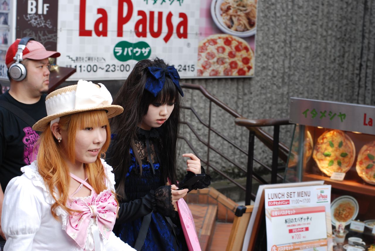 Two young women on a Harajuku street, one in a white and pink sweet Lolita coord with a straw bonnet and one in a navy and black Gothic Lolita dress