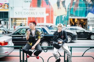 Two young men in Harajuku street style sitting on a rail, one with red dyed hair, printed shirt and black utility vest, the other in a black jacket with silver metallic cargo trousers