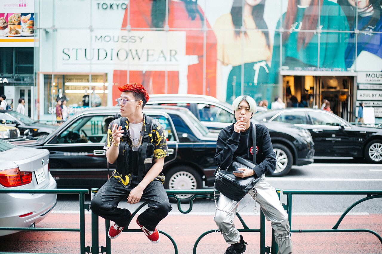 Two young men in Harajuku street style sitting on a rail, one with red dyed hair, printed shirt and black utility vest, the other in a black jacket with silver metallic cargo trousers