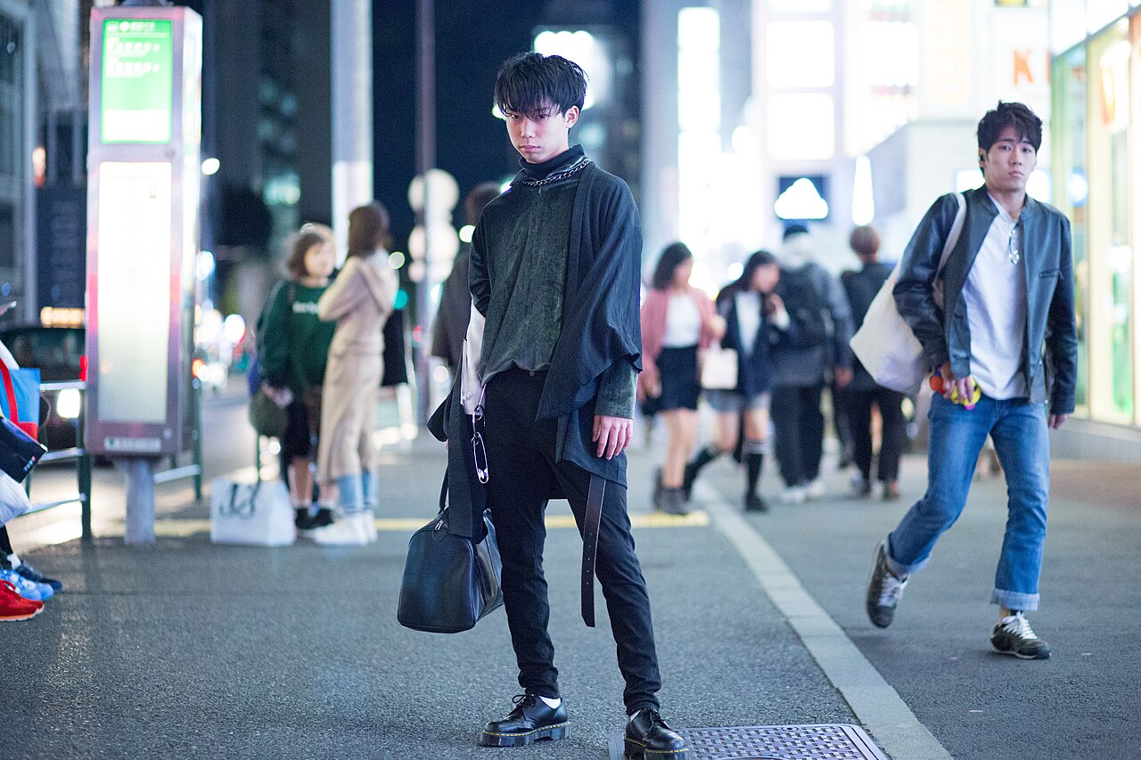 Young man walking a Tokyo street in a layered all-black Harajuku outfit with a draped asymmetric cardigan, tapered trousers, chunky platform derbies and a black tote bag