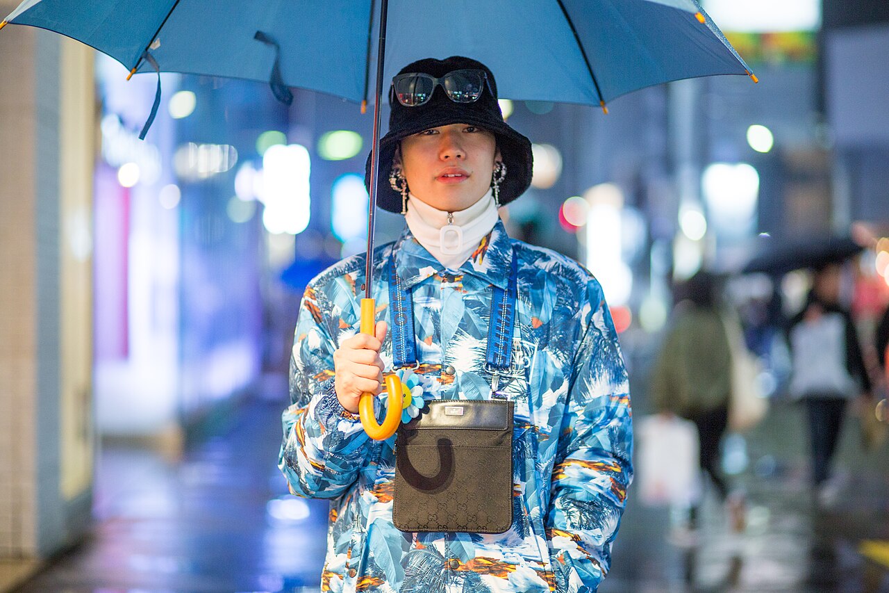 Young man in Harajuku wearing a blue and orange all-over print jacket, black bucket hat with sunglasses on the brim, white turtleneck and a small crossbody pouch