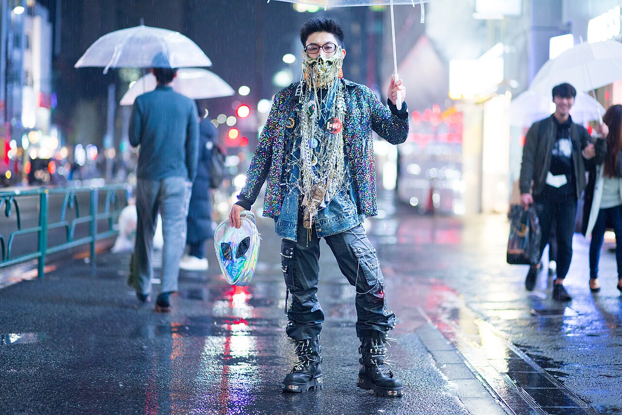 Man on a rainy Harajuku street at night in a glitter denim jacket covered in pins and chains, black cargo trousers and chunky buckled platform boots