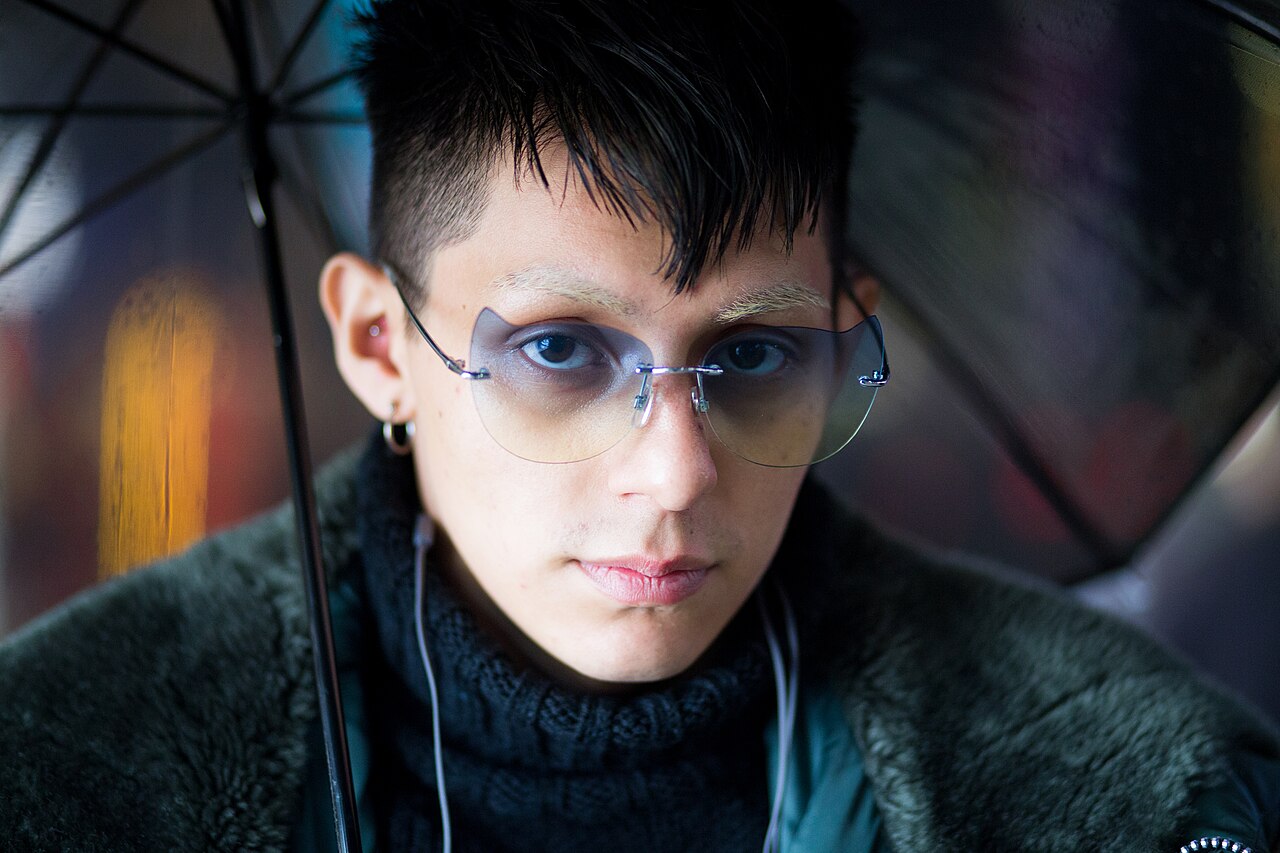 Close-up of a young man in Harajuku with an undercut, bleached eyebrows, blue tinted rimless glasses and a black knit turtleneck under a dark fur collar coat