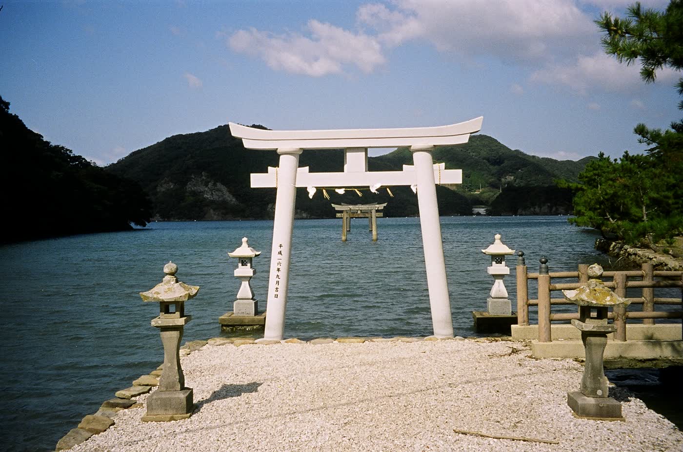 White Shinto torii gate standing at the water's edge with a second torii out in the sea and forested hills behind